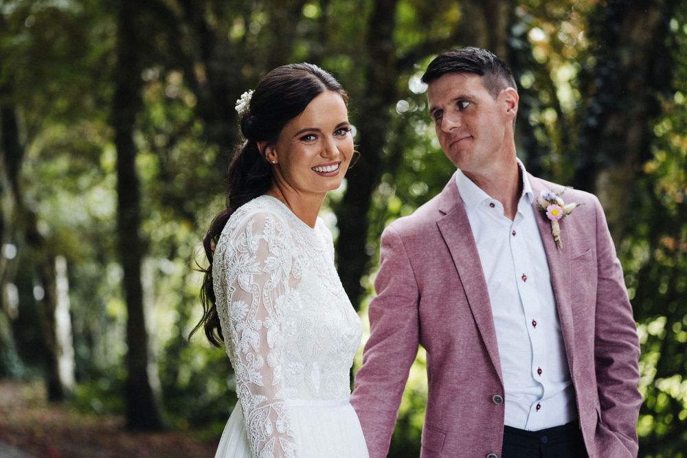 Wedding portrait in a lush woodland setting featuring elegant bridal attire and a distinctive pink jacket.