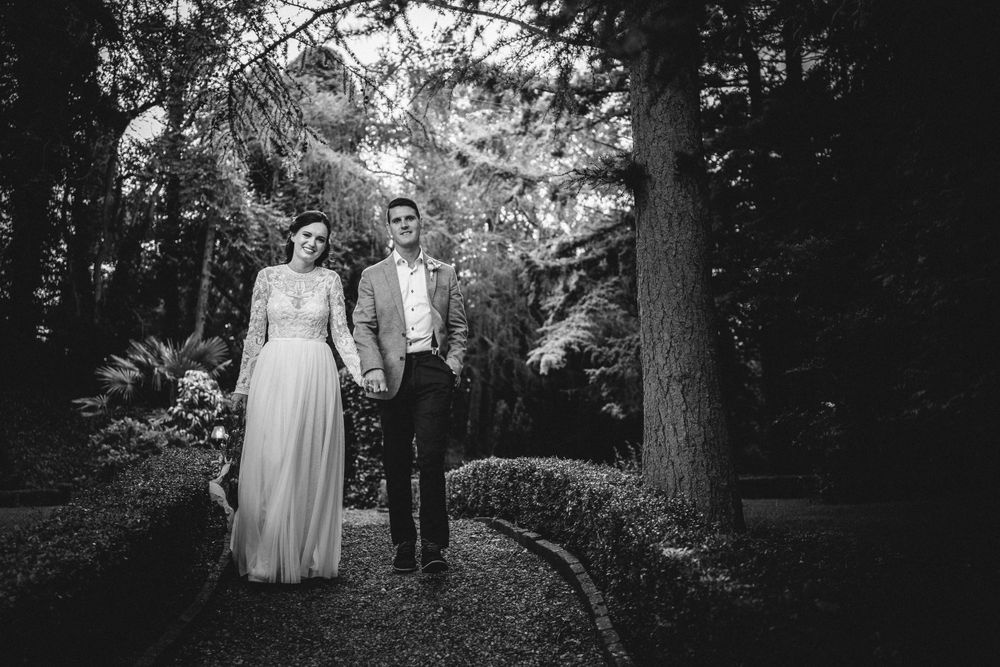 Black and white photograph of newlyweds walking along a garden path surrounded by tall trees.