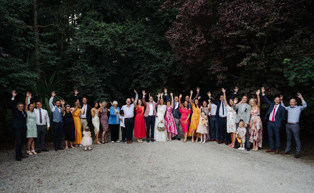 Large group wedding photo of guests celebrating outdoors against a backdrop of dark trees.