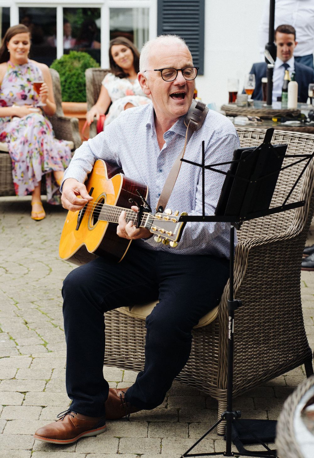 A guitarist with glasses plays an acoustic guitar while seated in wicker furniture at an outdoor gathering.