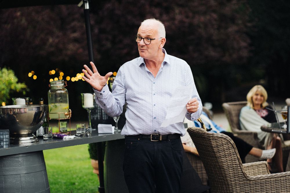 A speaker gestures while giving a speech at an outdoor garden party.
