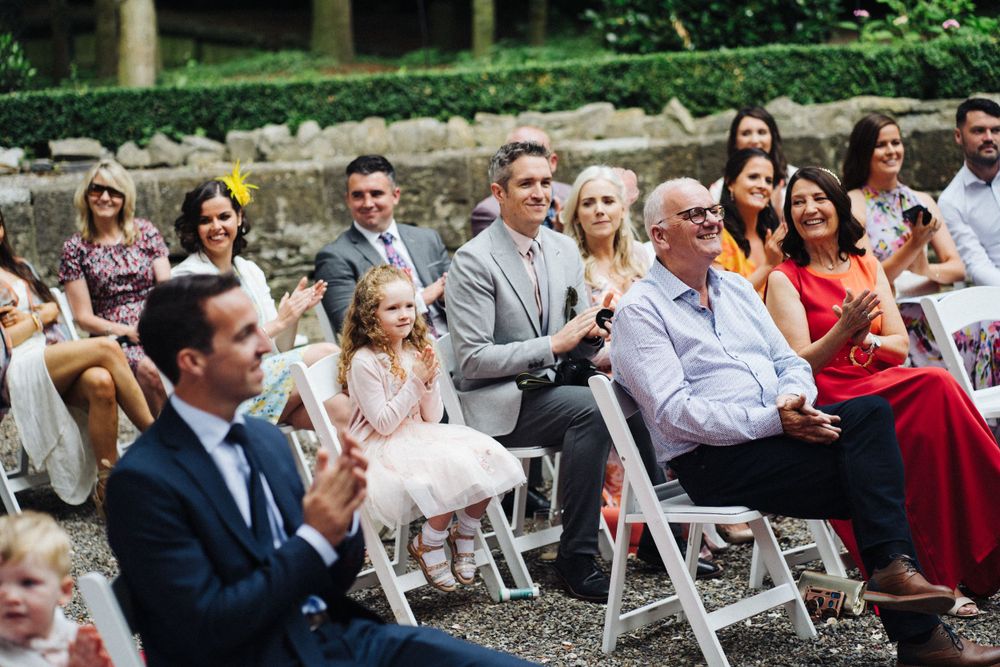 Outdoor wedding ceremony with guests seated on white chairs among trees.