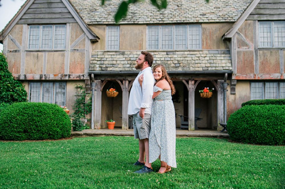 A sweet couple stands together in front of a charming Tudor-style house with manicured hedges.