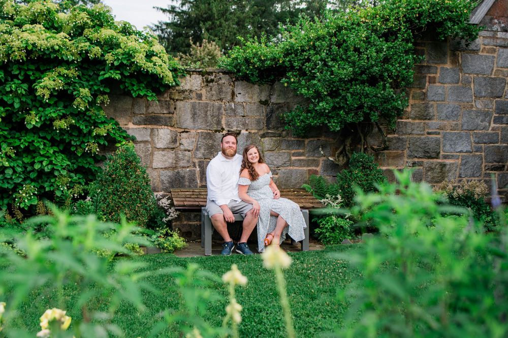 A couple relaxes on a wooden bench in a serene garden setting with stone walls and green foliage.