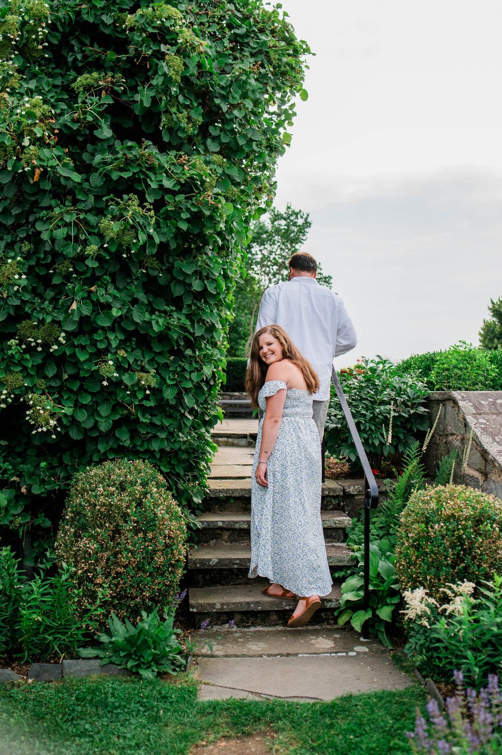 A couple ascends stone garden steps flanked by manicured shrubs and climbing vines.