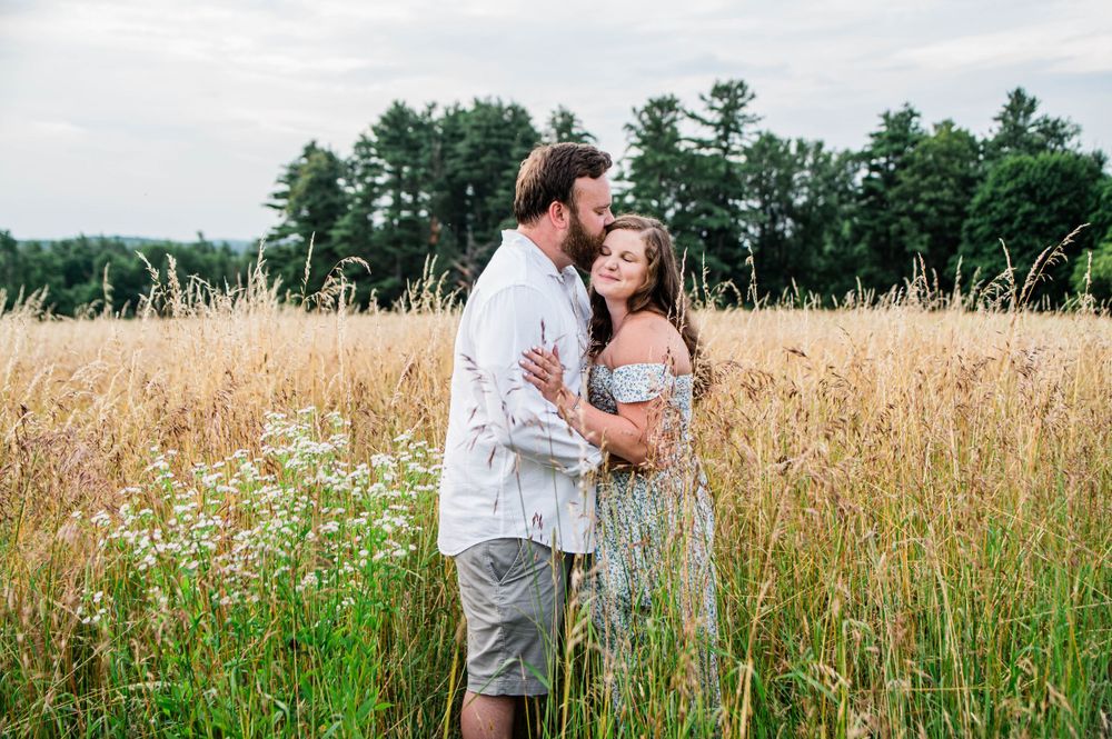 A romantic portrait of a couple embracing in a sun-lit field of tall golden grass with evergreen trees in the background.