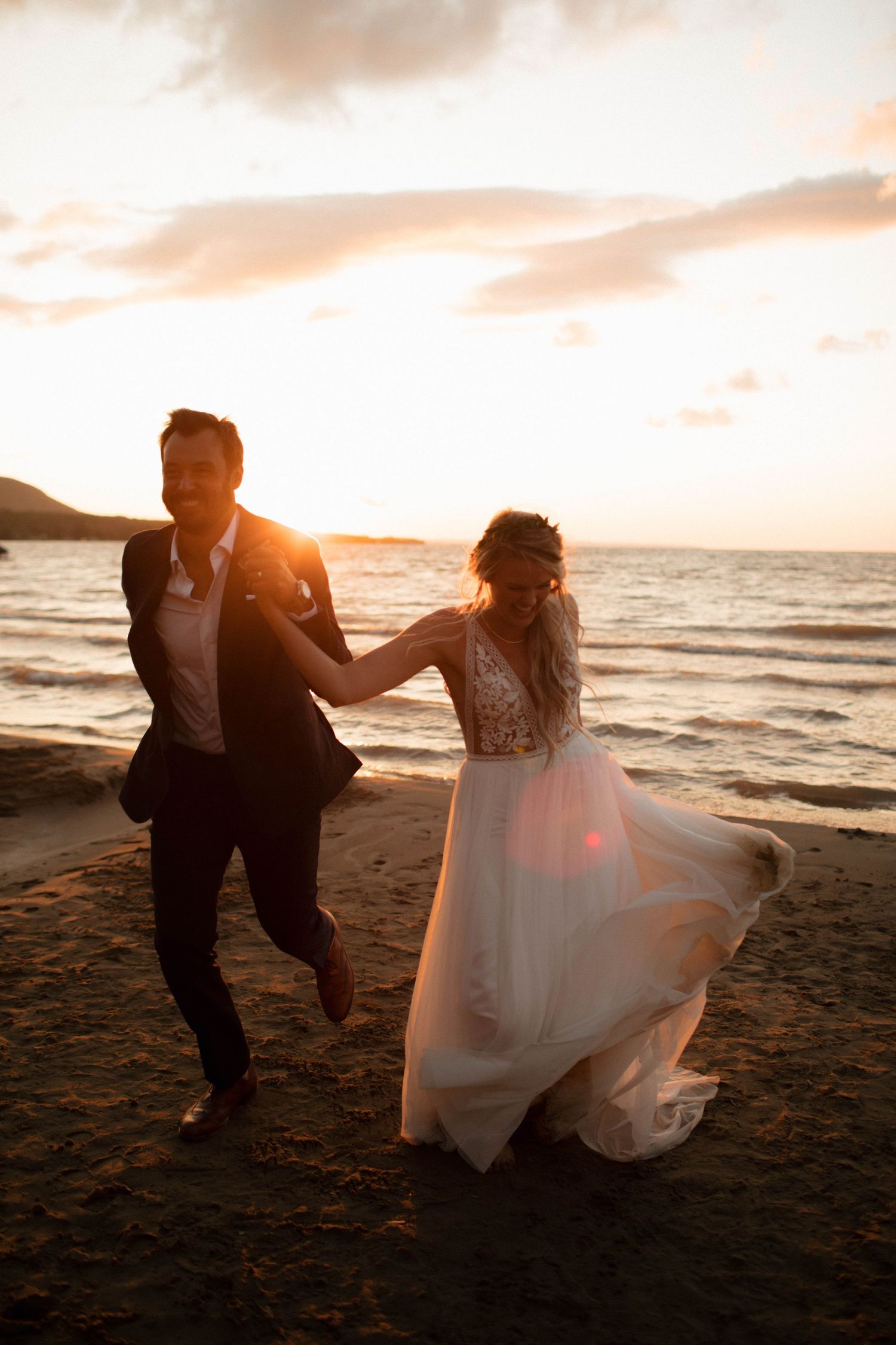 Couple dancing and laughing on a beach at sunset in wedding attire.