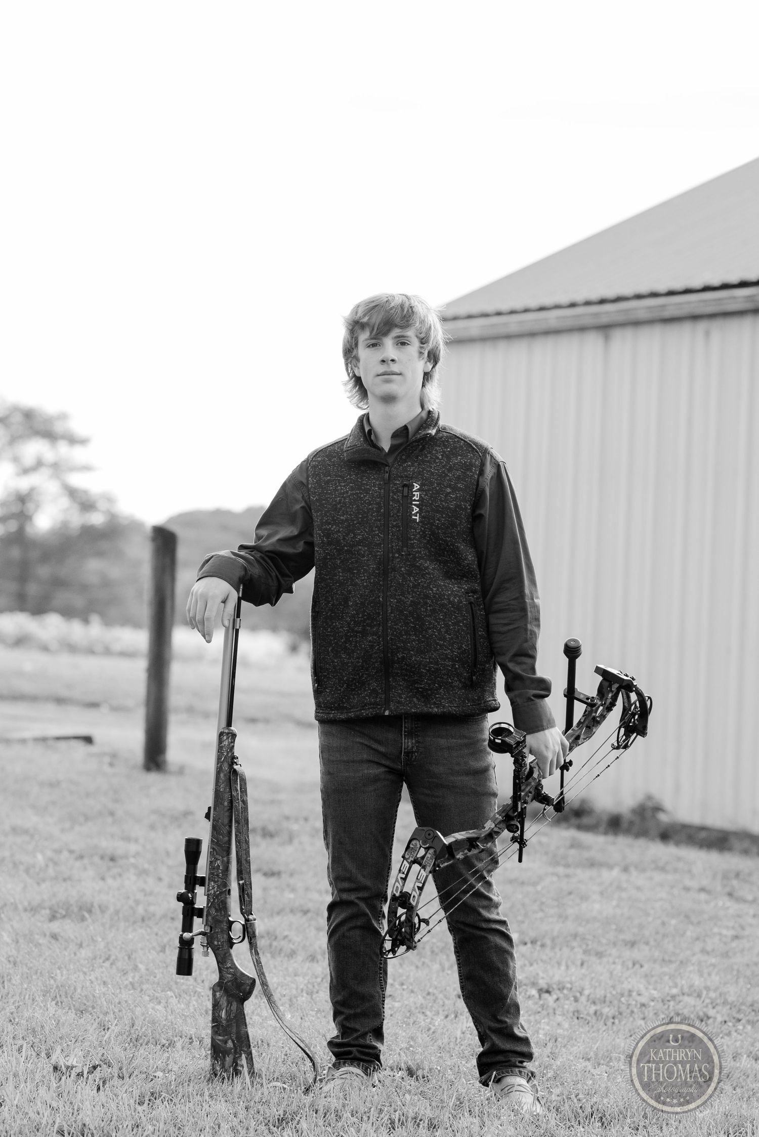 Black and white photo showing person with archery and hunting equipment by metal building.