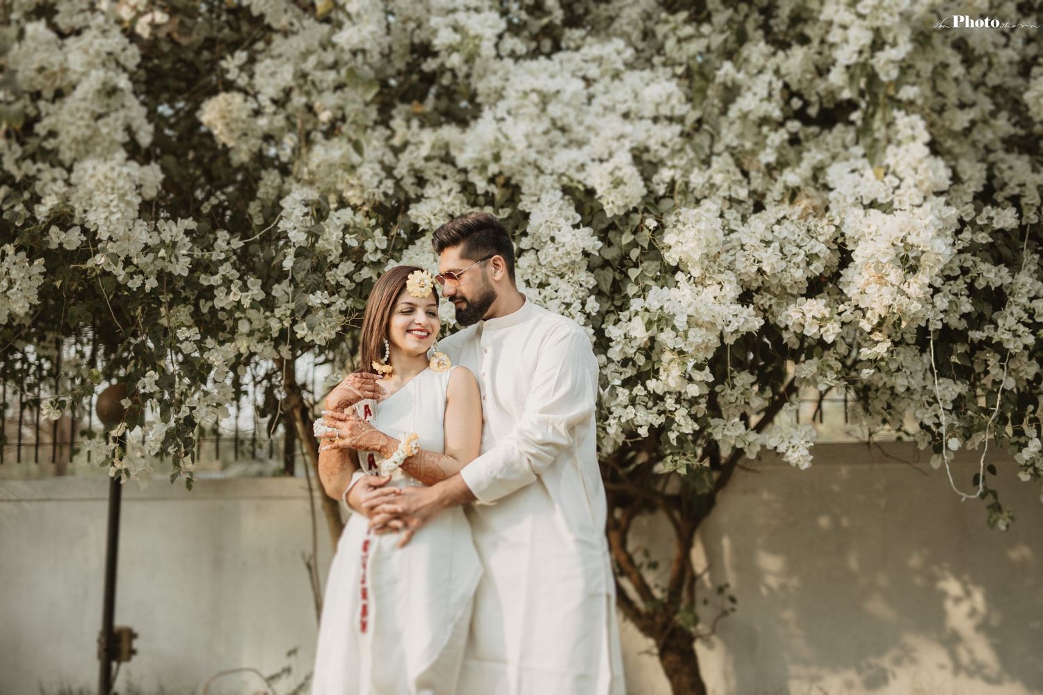 Romantic wedding couple embraces under blooming white cherry blossom trees in an outdoor garden setting.