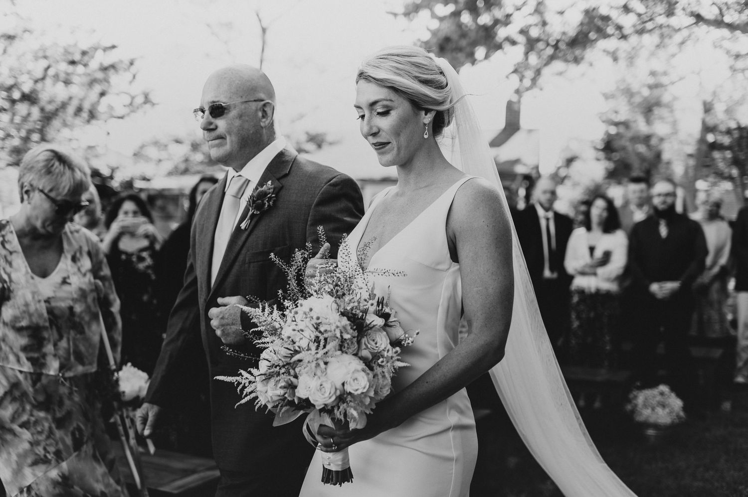 Black and white photo of a wedding ceremony under trees with guests watching.