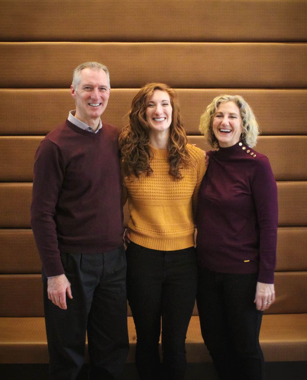 Three people wearing coordinated sweaters pose against wooden wall background.