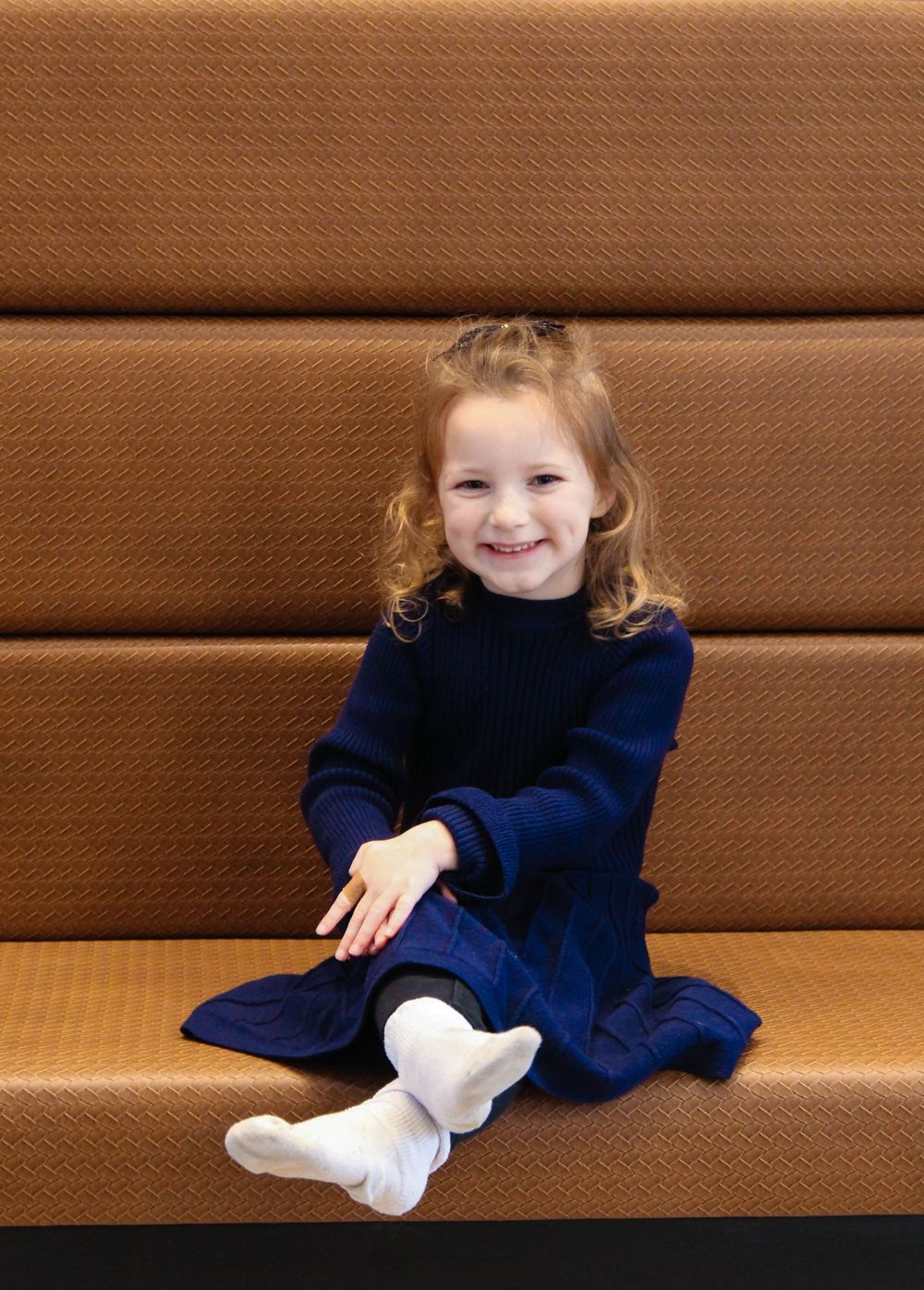 Young child in navy dress sitting on brown cushioned bench with bright smile.