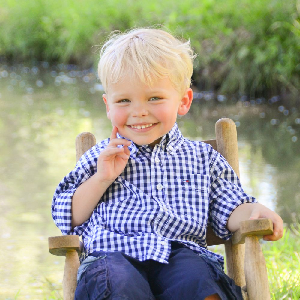 A young child in a blue checkered shirt sitting on a wooden chair outdoors.