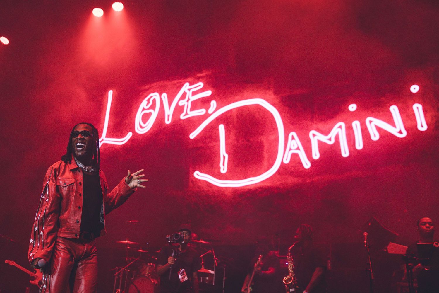 Concert stage bathed in red light with neon 'Love Damini' sign and performer in action.