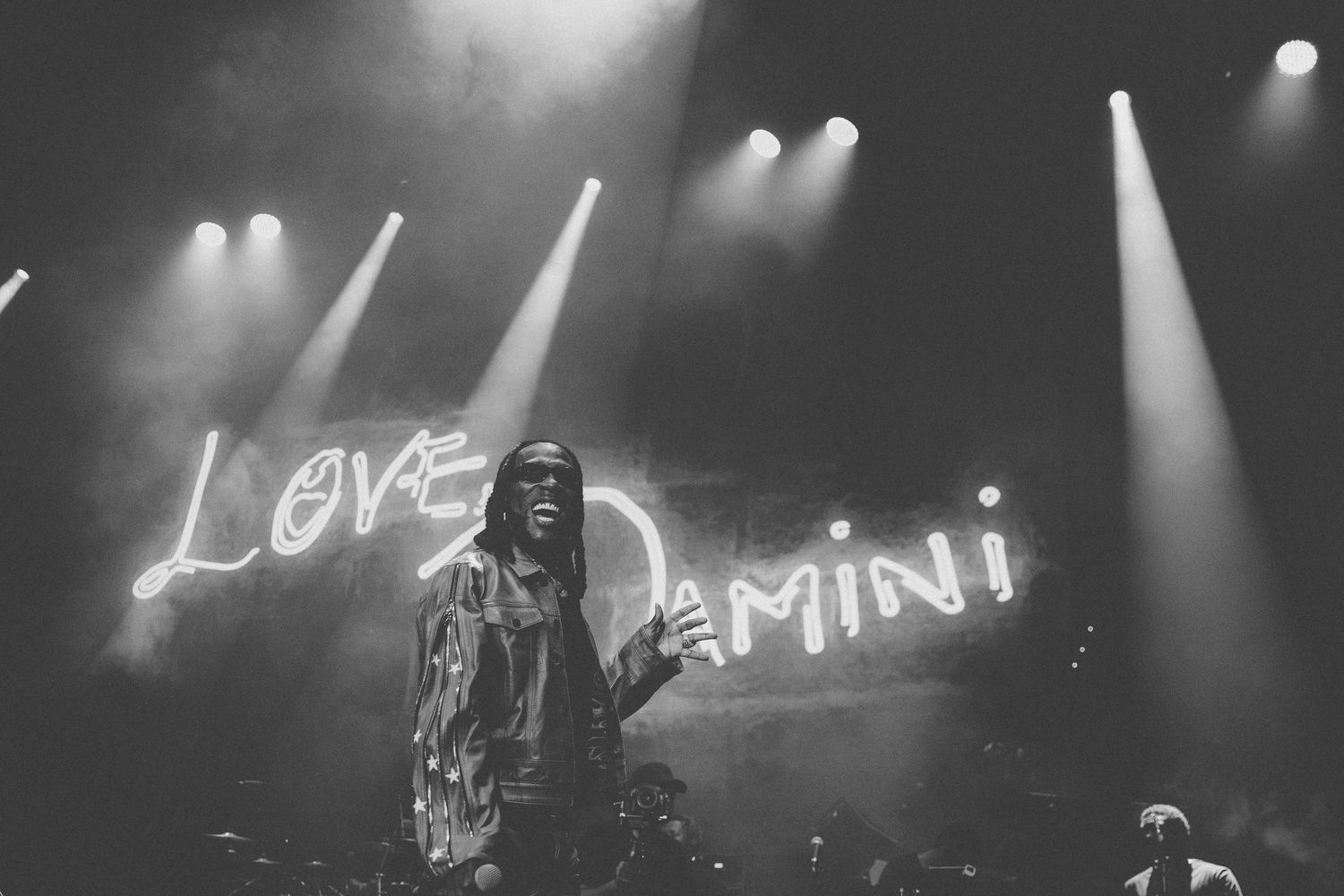 Black and white concert photo showing performer in front of illuminated 'Love Damini' sign.
