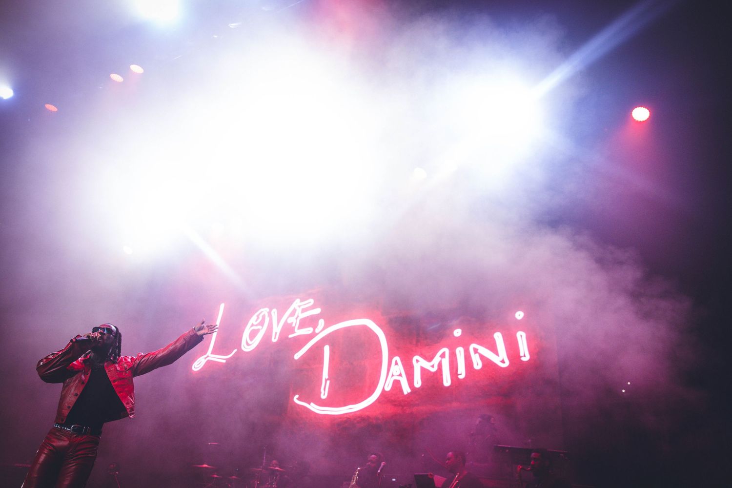 Concert stage with bright neon 'Love Damini' sign glowing through atmospheric smoke and lighting.