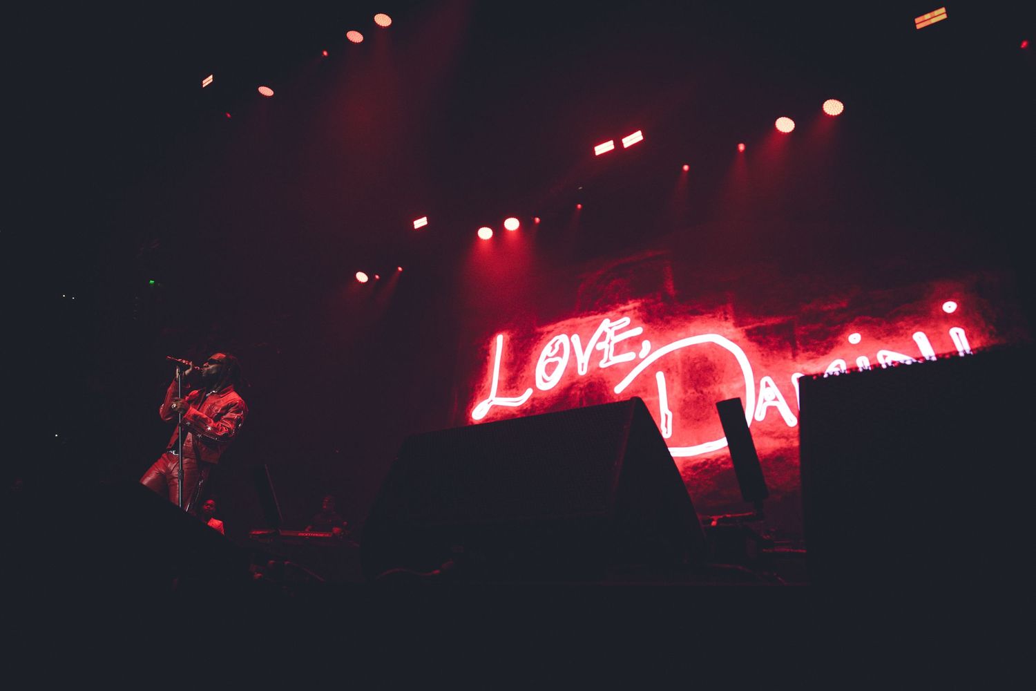 Dark concert venue with 'Love Dam' neon sign glowing red against black background.