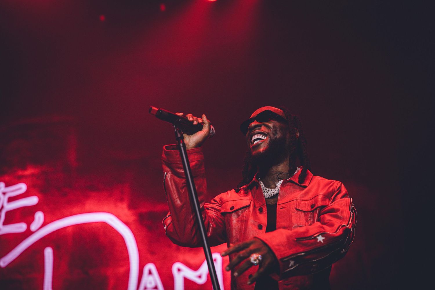A performer in a red jacket sings on stage with dramatic red neon lighting in the background.