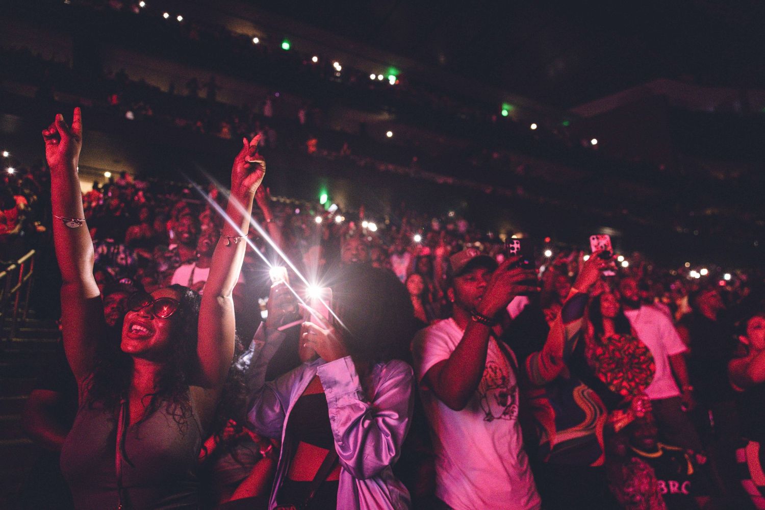 Concert crowd with raised hands illuminated by red lights and stage beams during nighttime performance.