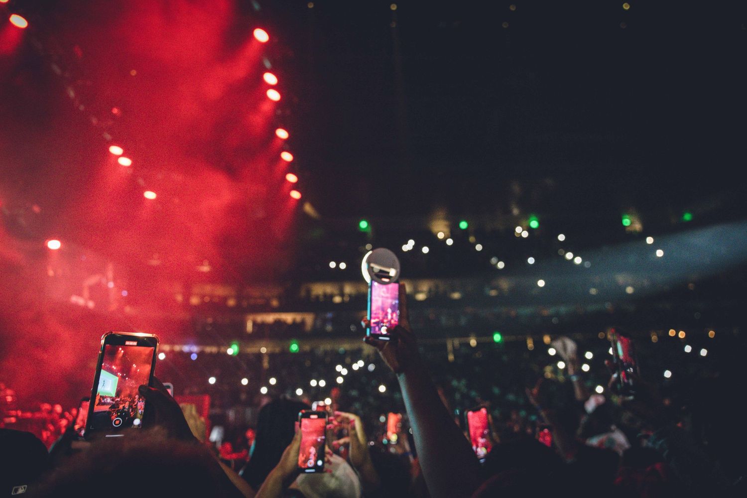 Concert crowd holds up phones creating a sea of lights while performer appears on stage in red lighting.