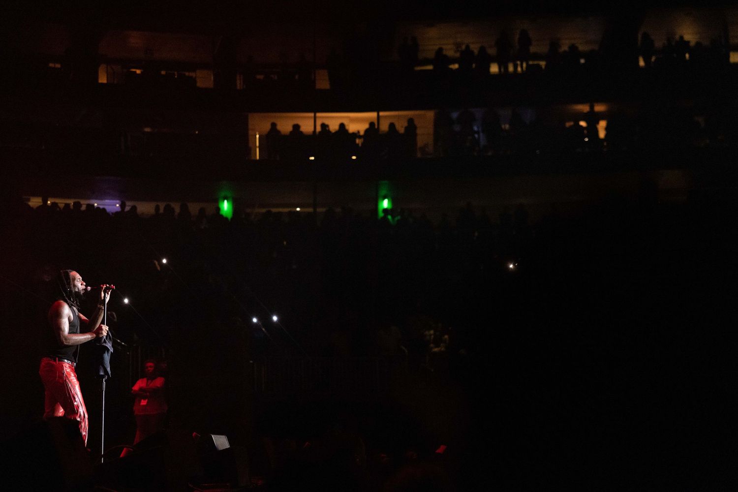 A dimly lit concert venue with a performer on stage surrounded by dark silhouettes.