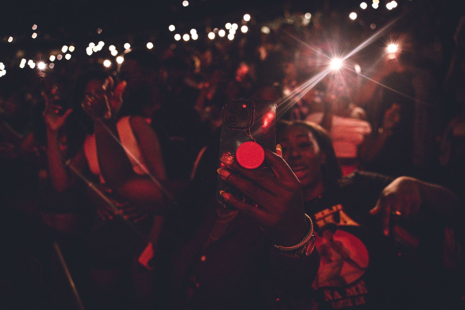 Concert crowd illuminated by red stage lights with glowing cell phones raised in the darkness.