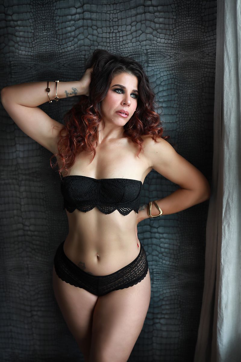 Artistic boudoir photo of model in black lace lingerie against dark textured wall.