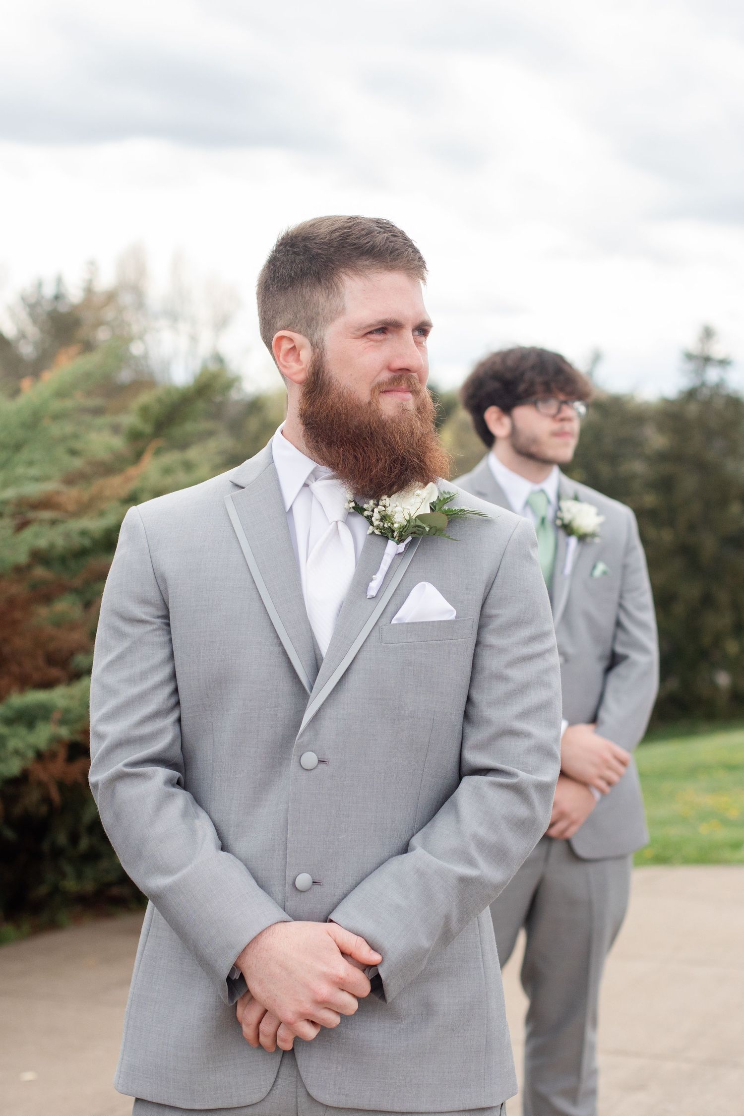 Groomsmen in light gray suits and white ties stand together outdoors during a wedding.