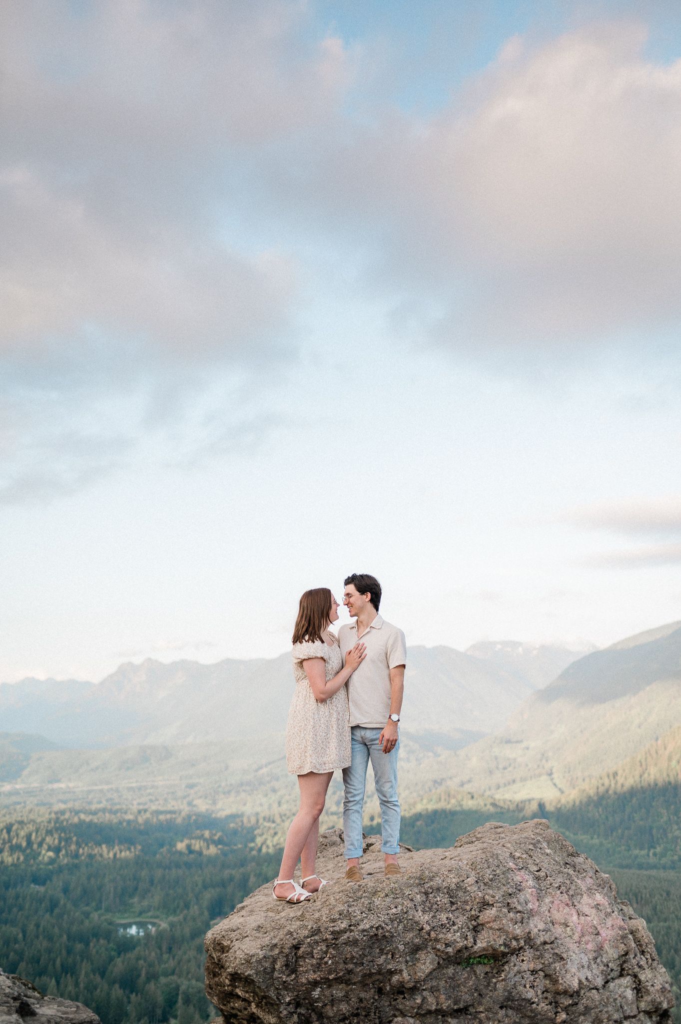 A couple stands together on a rocky mountain overlook with a scenic forest valley and mountain range in the background with dramatic clouds
