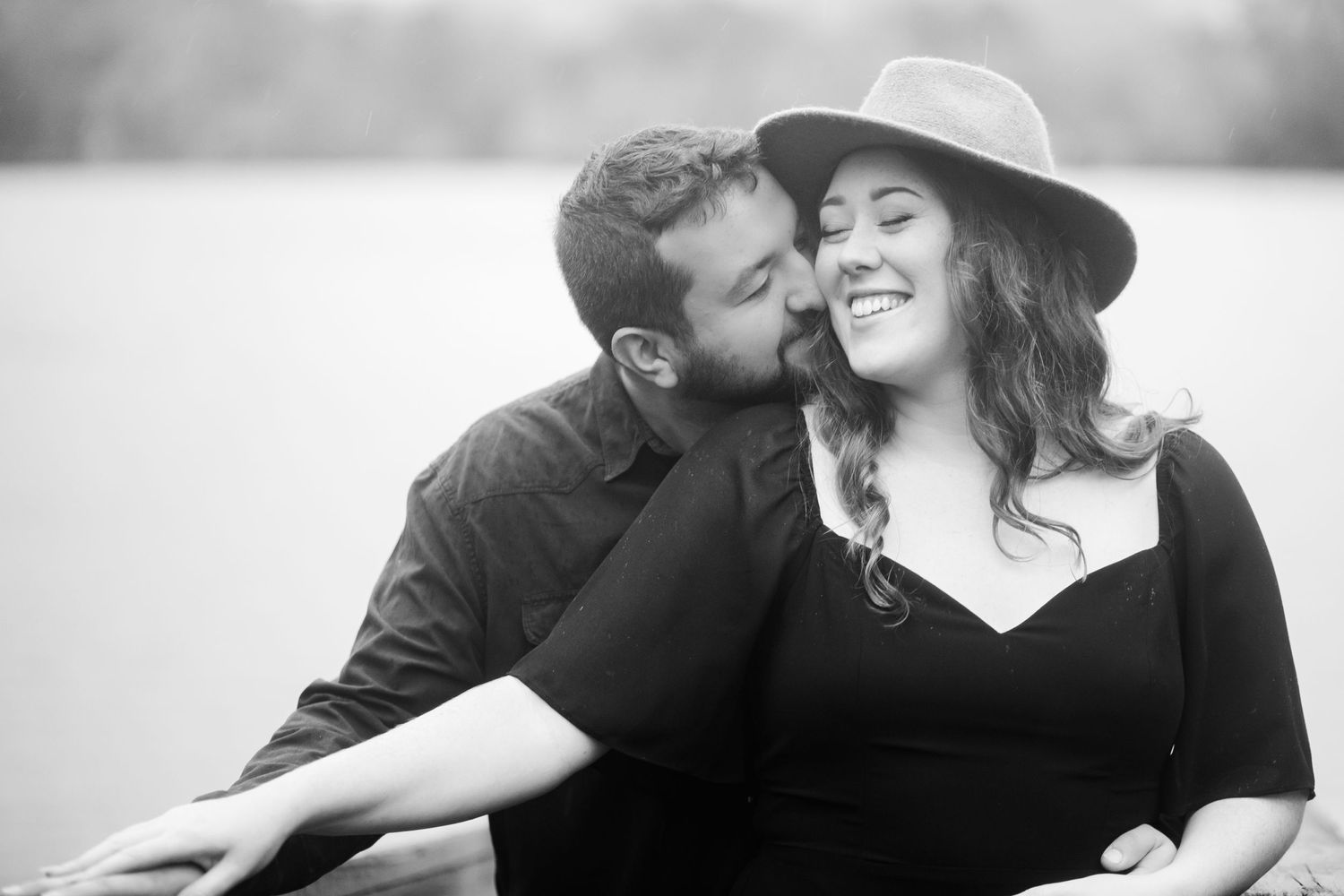 Black and white photo of couple sharing affectionate moment outdoors with lake backdrop.