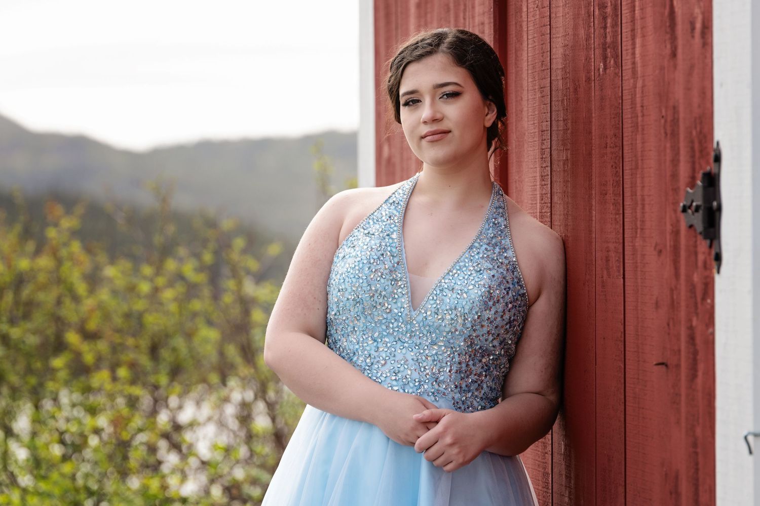 Young woman in sparkly blue prom dress poses by red barn door with mountains in background.