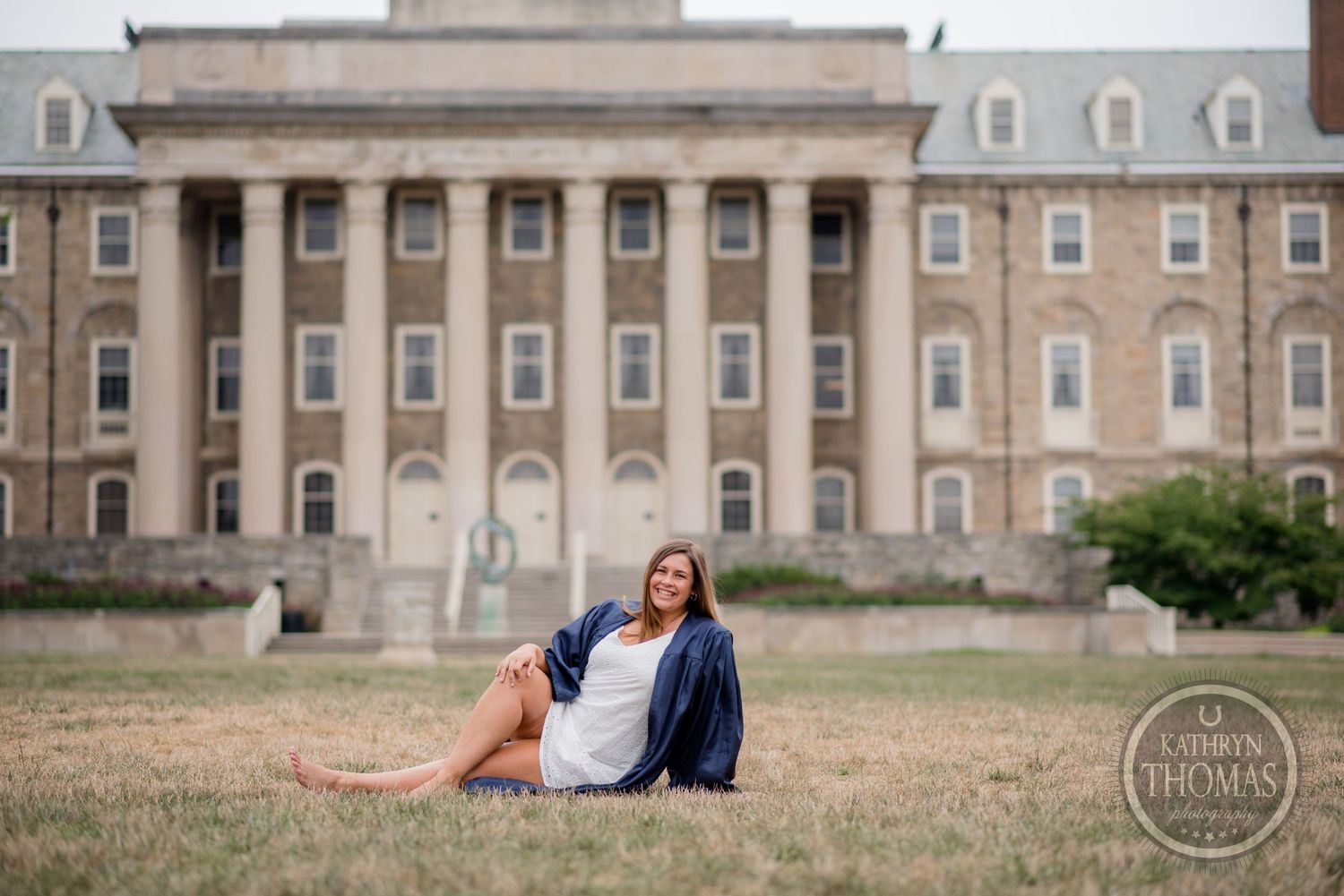 Graduate in blue cap and gown sits on grass in front of grand collegiate building facade.