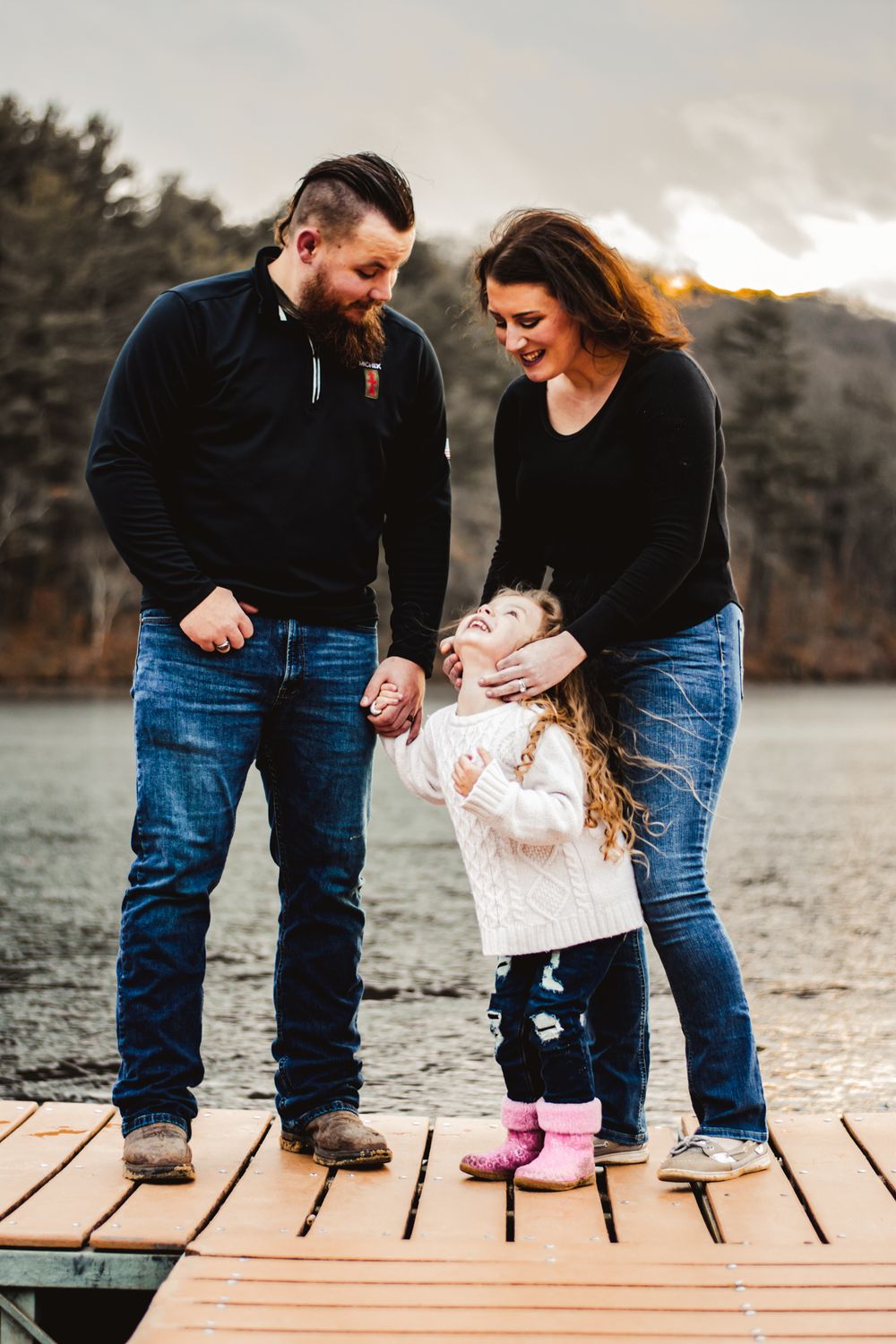 A family shares laughter while standing together on a lakeside dock during sunset.
