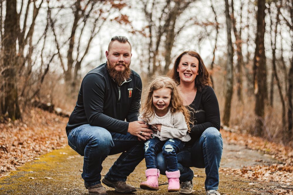 Family portrait on autumn path with fallen leaves and bare trees in background.
