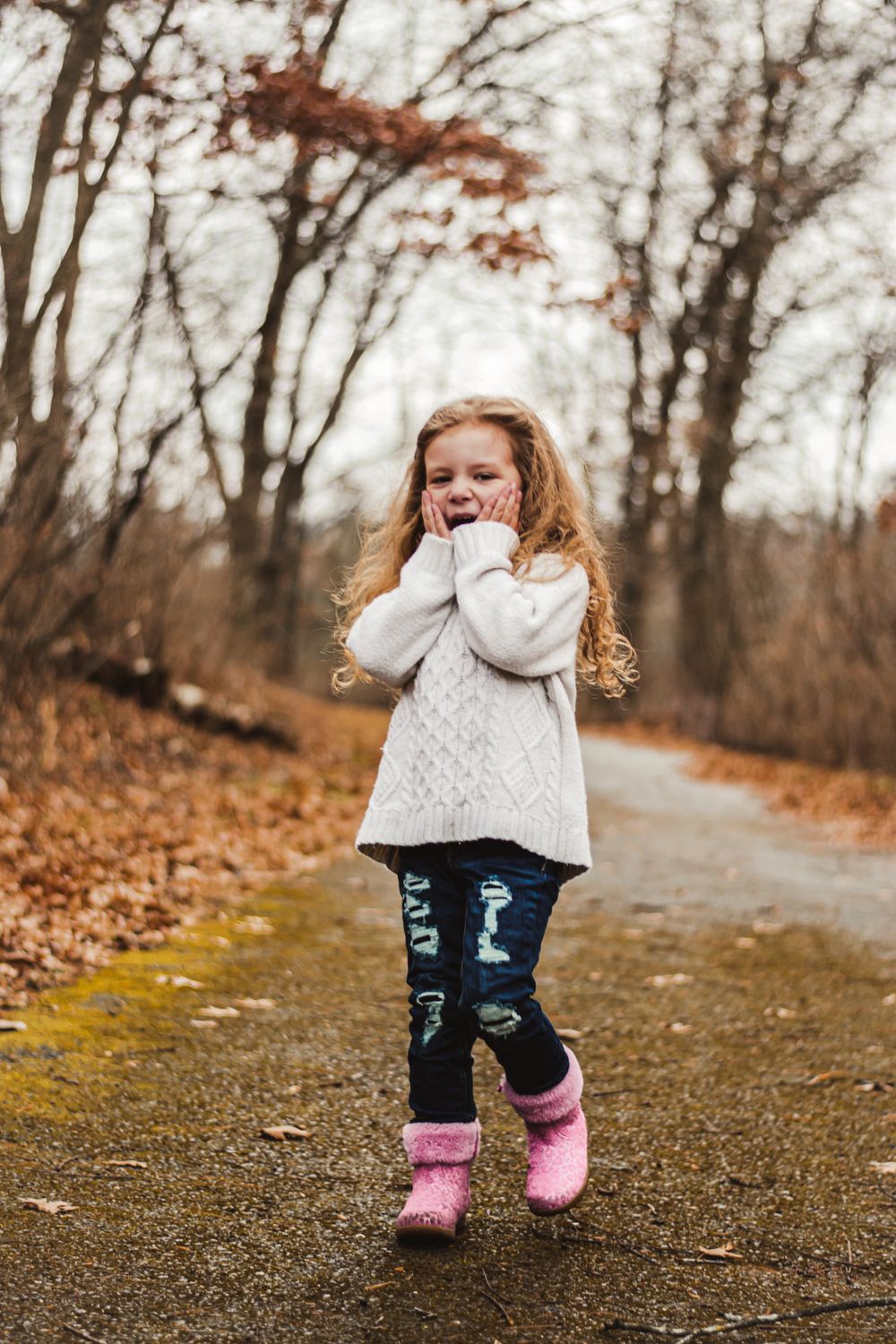 Child in white sweater and pink boots expressing joy on autumn forest path.