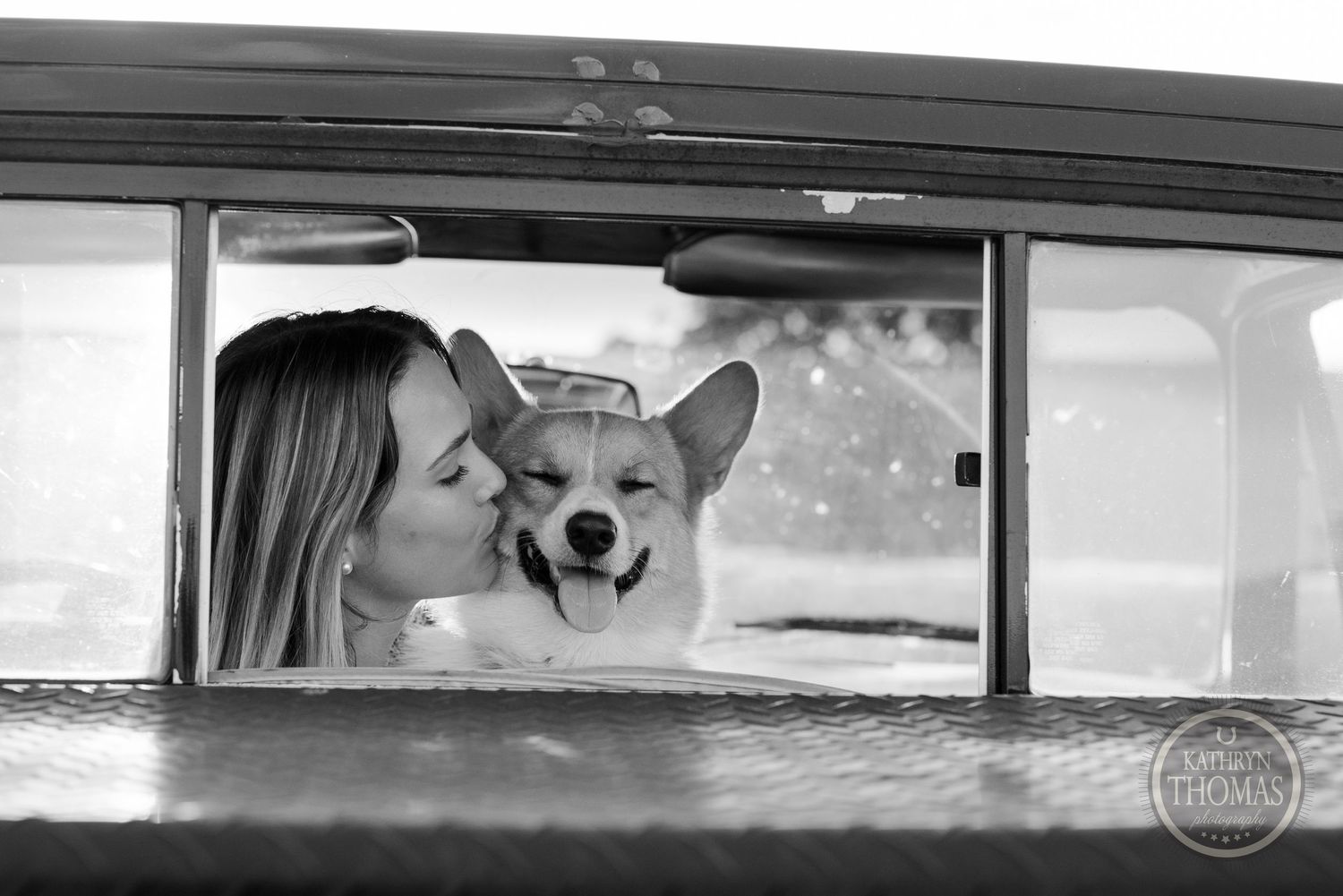 A black and white photo of a smiling Corgi dog looking through a vintage car window.