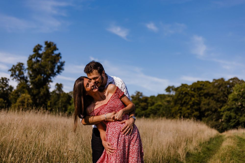 A couple embracing in a tall grass field on a sunny day with trees in the background.