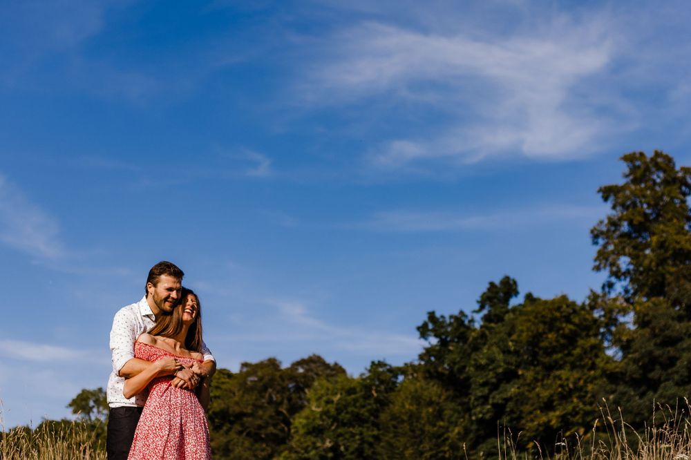 Couple embracing in a sunny field with blue sky and trees in the background.