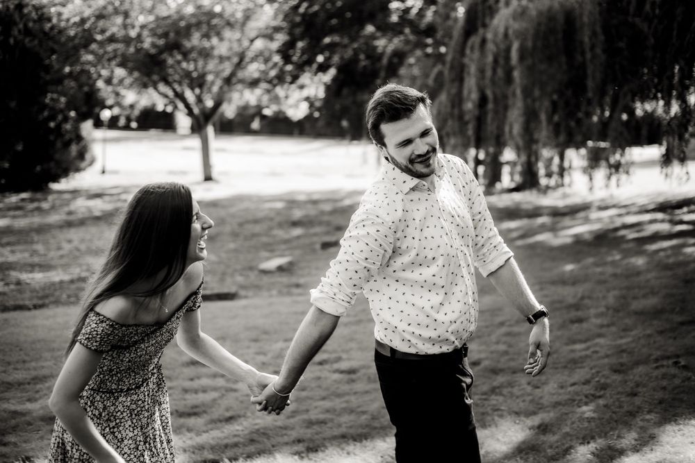 Black and white photo of a couple holding hands and walking in a park with willow trees.