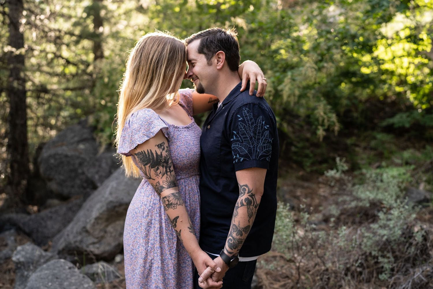 A romantic couple embraces in a sunlit forest setting with the woman wearing a flowing lavender dress.