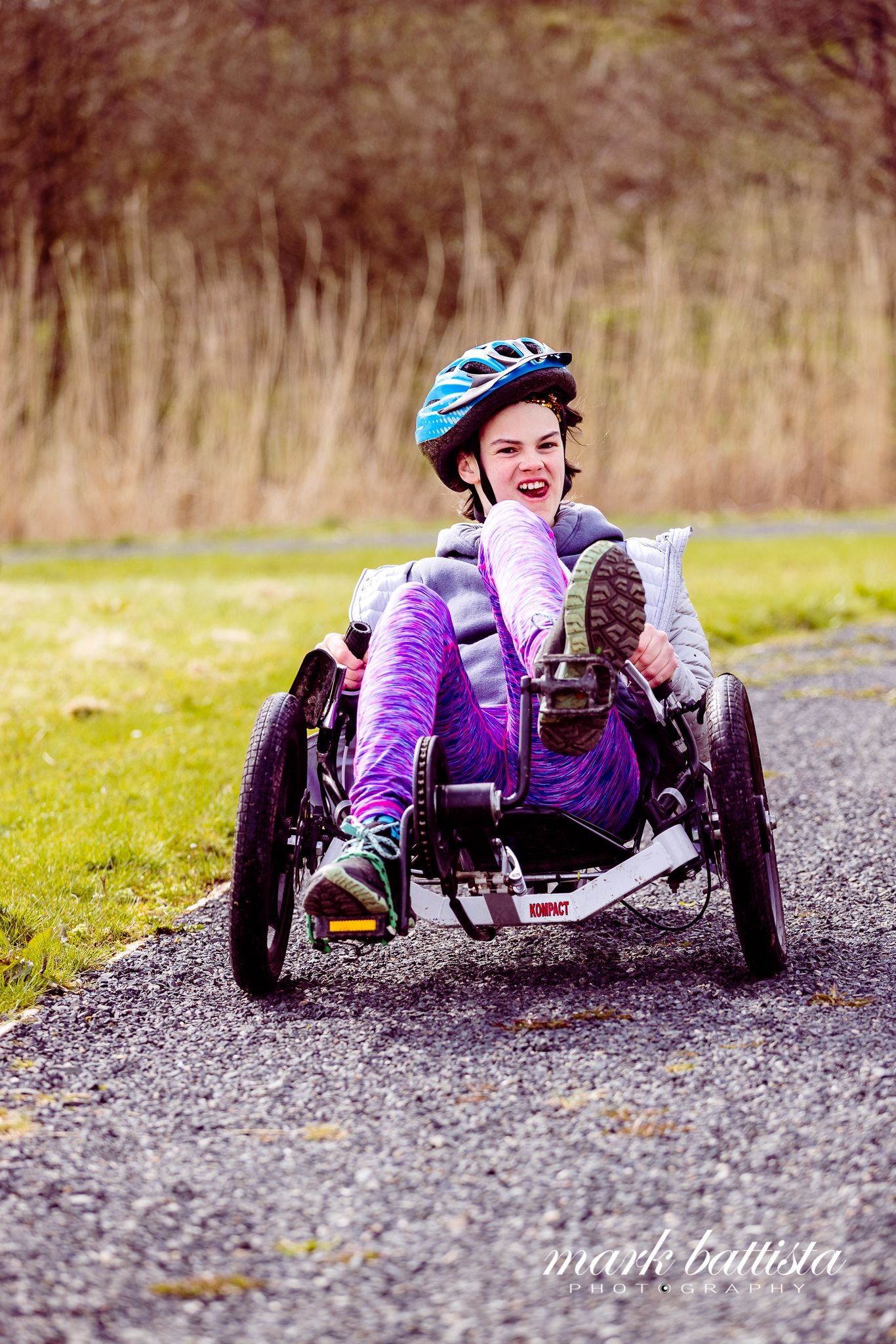 Adaptive athlete riding a recumbent trike on a trail wearing purple and blue gear.