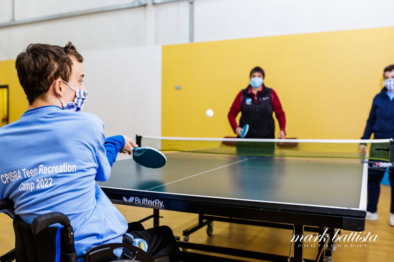 Players participating in an adaptive table tennis match in a gym with yellow walls.