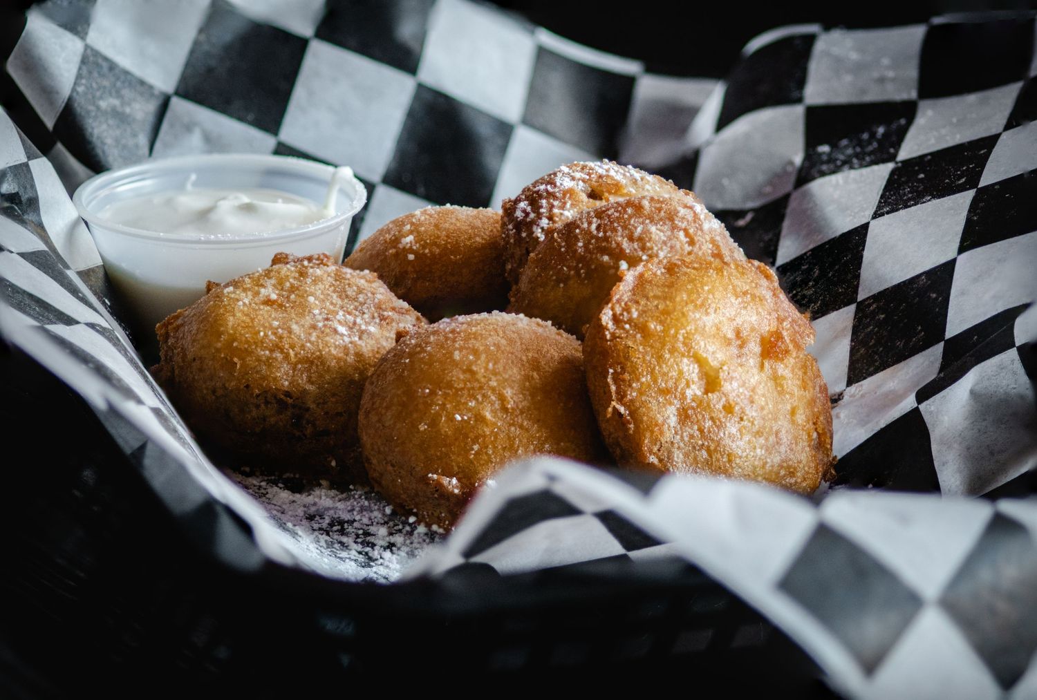 Golden fried donut holes dusted with powdered sugar served in a checkered basket with dipping sauce.