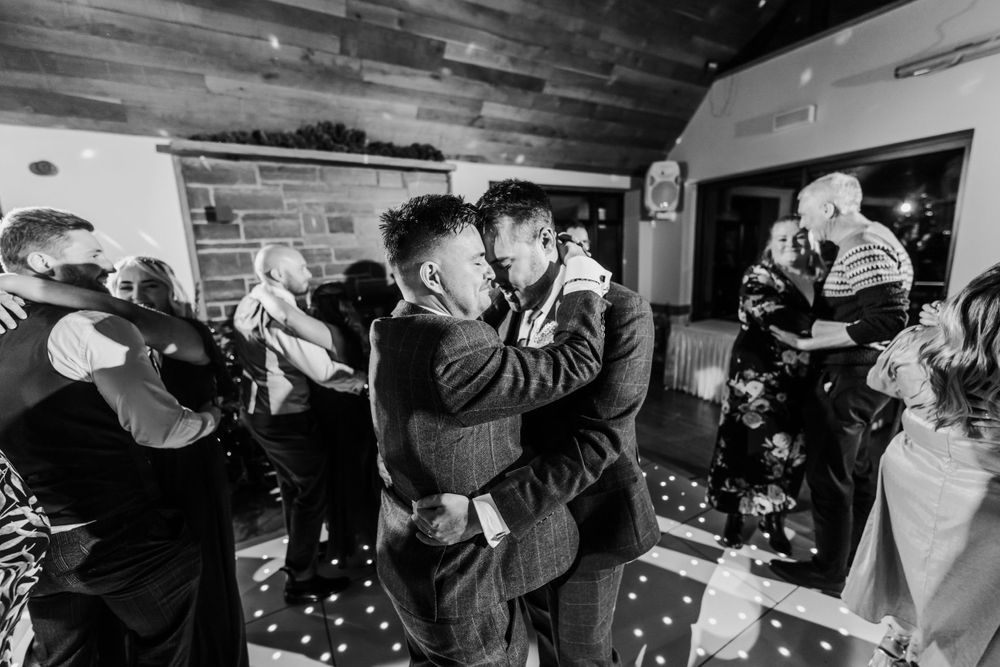 A black and white candid shot of wedding guests dancing and celebrating on a wooden floor.