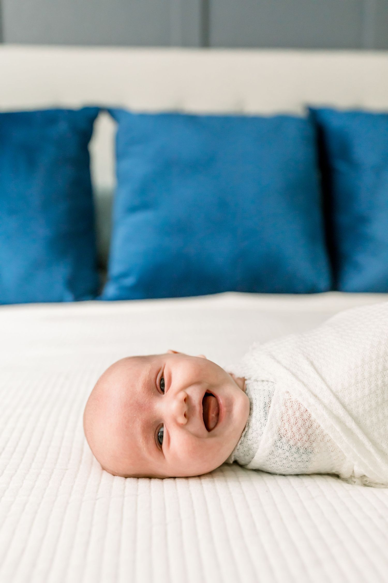 A baby in a white swaddle smiles while lying on a white bed with blue accent pillows.