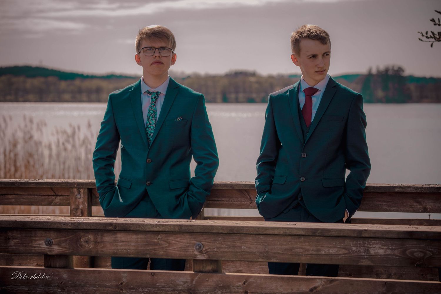 Two young men in matching teal suits pose on a wooden dock by a lake.