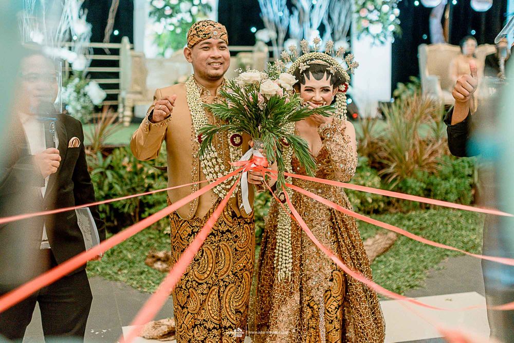 A couple wearing traditional Javanese wedding attire celebrates with festive red ribbons.