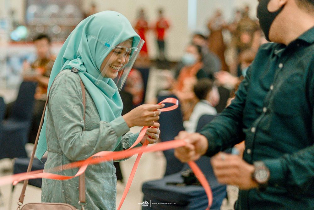 A woman in a grey hijab smiles while holding a red ribbon during a wedding game.