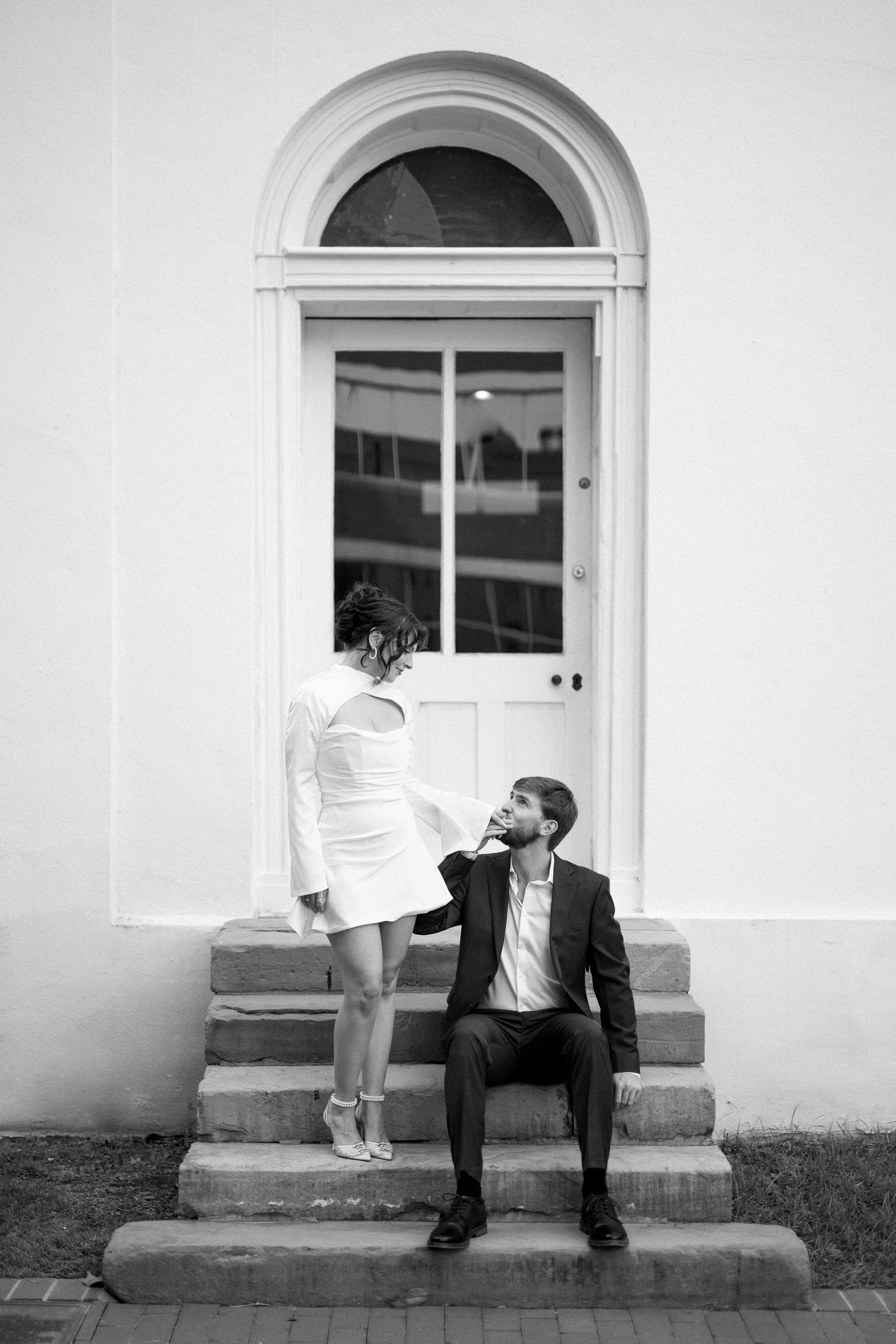 Black and white photo of a couple on stone steps with an elegant arched doorway backdrop.