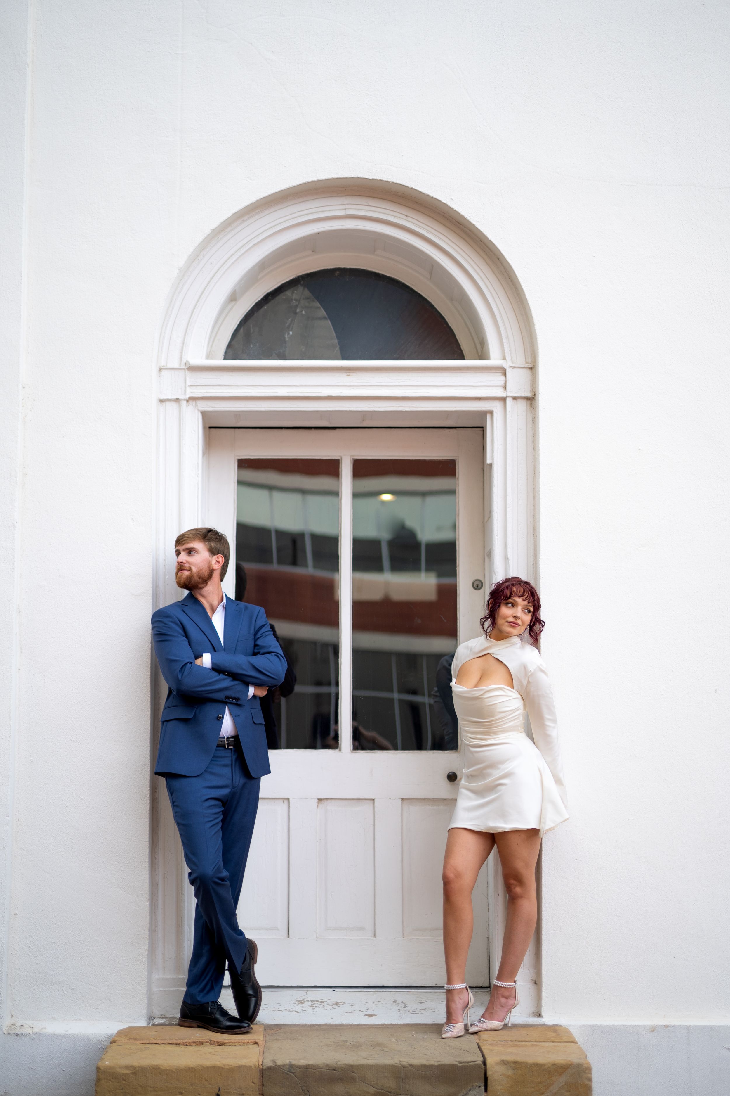 Couple in formal attire interacts playfully by white building entrance.